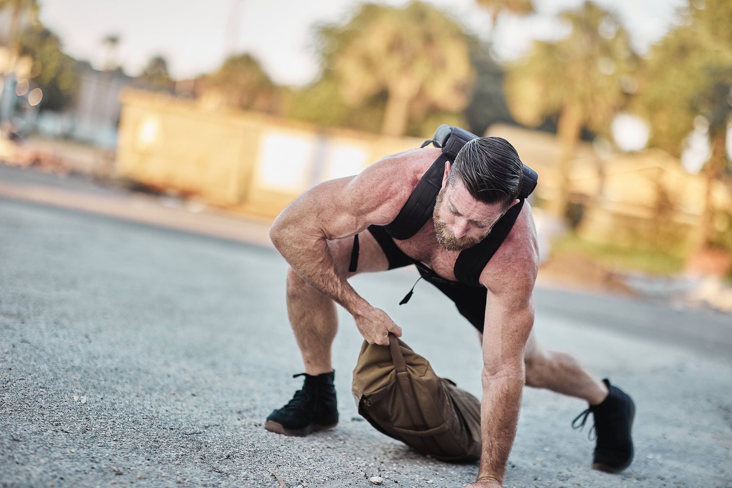 A muscular man in workout gear lifts a sandbag outdoors on a sunny day, wearing the RPC Long + 30LB (Long) Plate Bundle weight vest for added resistance.