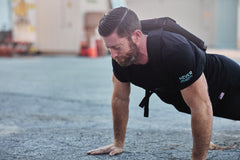A bearded man does push-ups outdoors on pavement while wearing a black shirt and the RPC Long + 30LB (Long) Plate Bundle weight vest for added resistance.