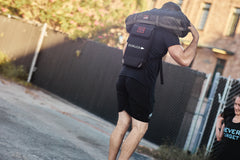 A man in athletic wear lifts the RPC Long + 30LB (Long) Plate Bundle outdoors, highlighting its ergonomic lumbar support, while another person observes in the background.