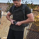 A man fastens a buckle on his black backpack, equipped with the Padded Hip Belt, while standing by a chain-link fence outdoors.