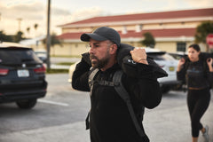 A man in black clothing and cap carries a RPC 3.0 + 10lb Ruck Sandplate on his shoulders during an outdoor weighted training session.