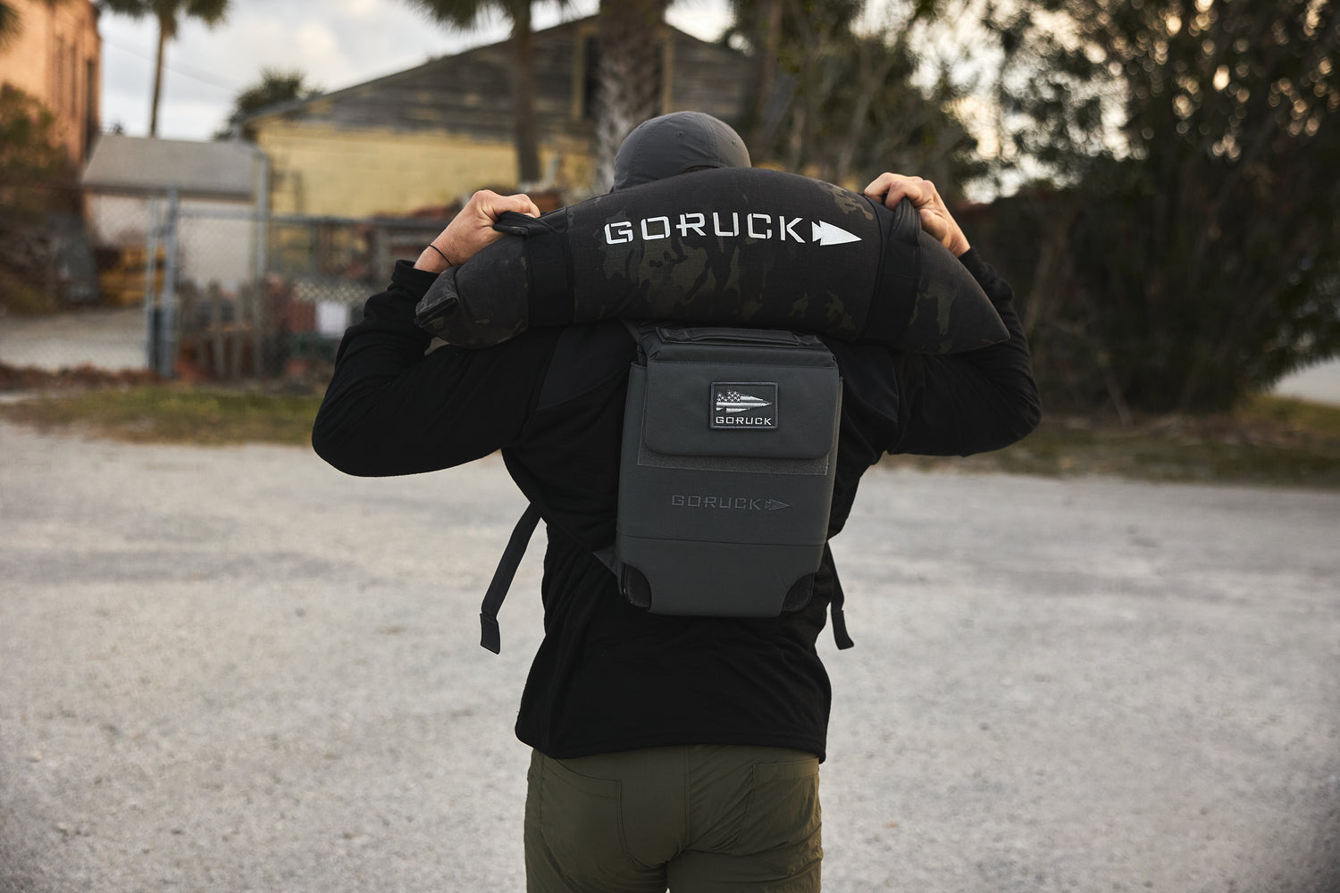 Outdoors, a person lifts the RPC 3.0 + 10lb Ruck Sandplate over their shoulders and wears a RPC 3.0 backpack with a sandplate for weighted training.