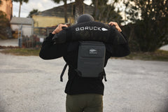Outdoors, a person lifts the RPC 3.0 + 10lb Ruck Sandplate over their shoulders and wears a RPC 3.0 backpack with a sandplate for weighted training.