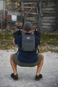 A man squats outdoors, holding a 10lb Ruck Sandplate at chest level for weighted training while wearing the RPC 3.0 backpack.