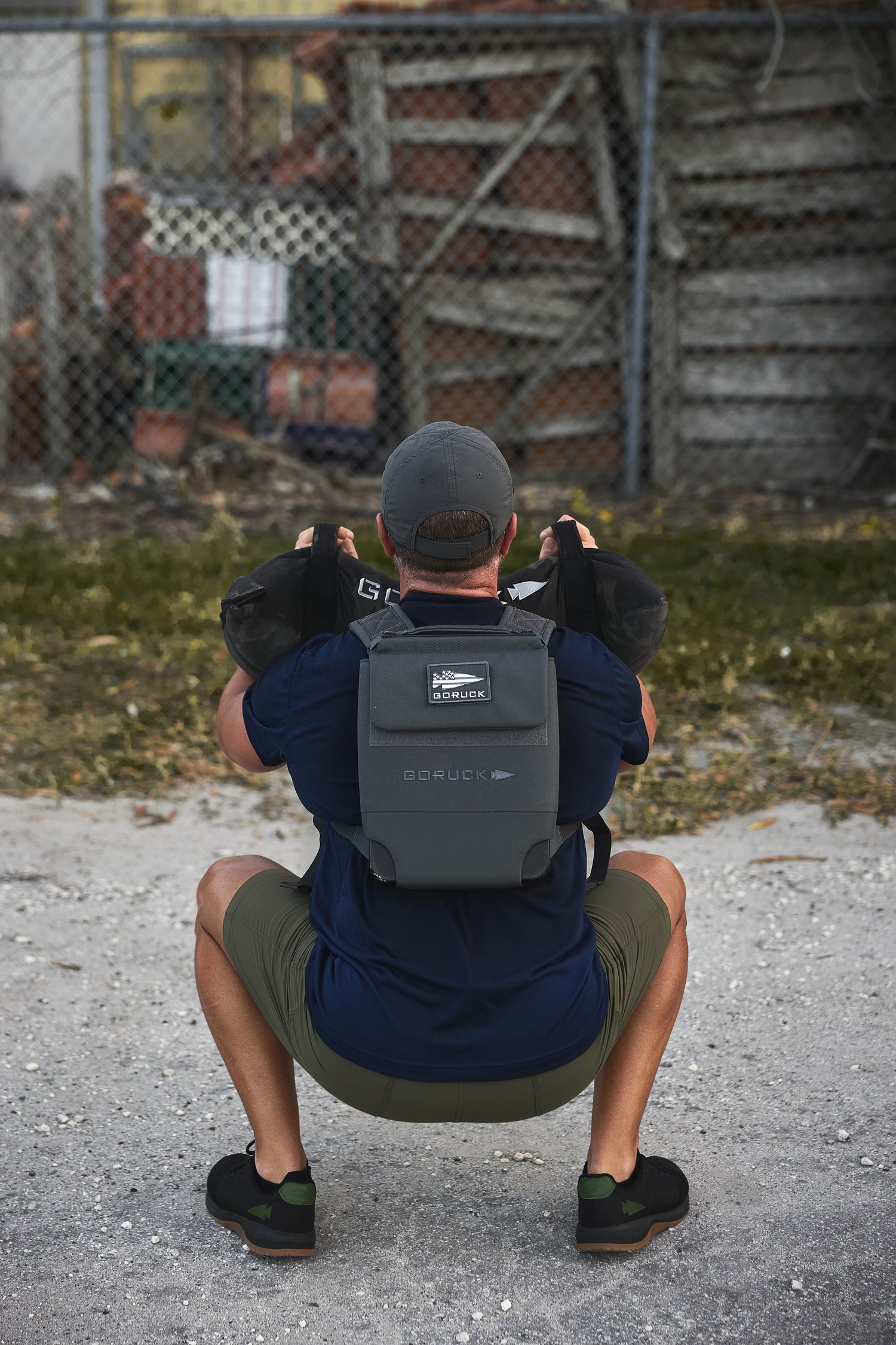A man outdoors squats near a chain-link fence and wooden crates, wearing the RPC 3.0 vest with a 10lb Ruck Sandplate and holding a sandbag.