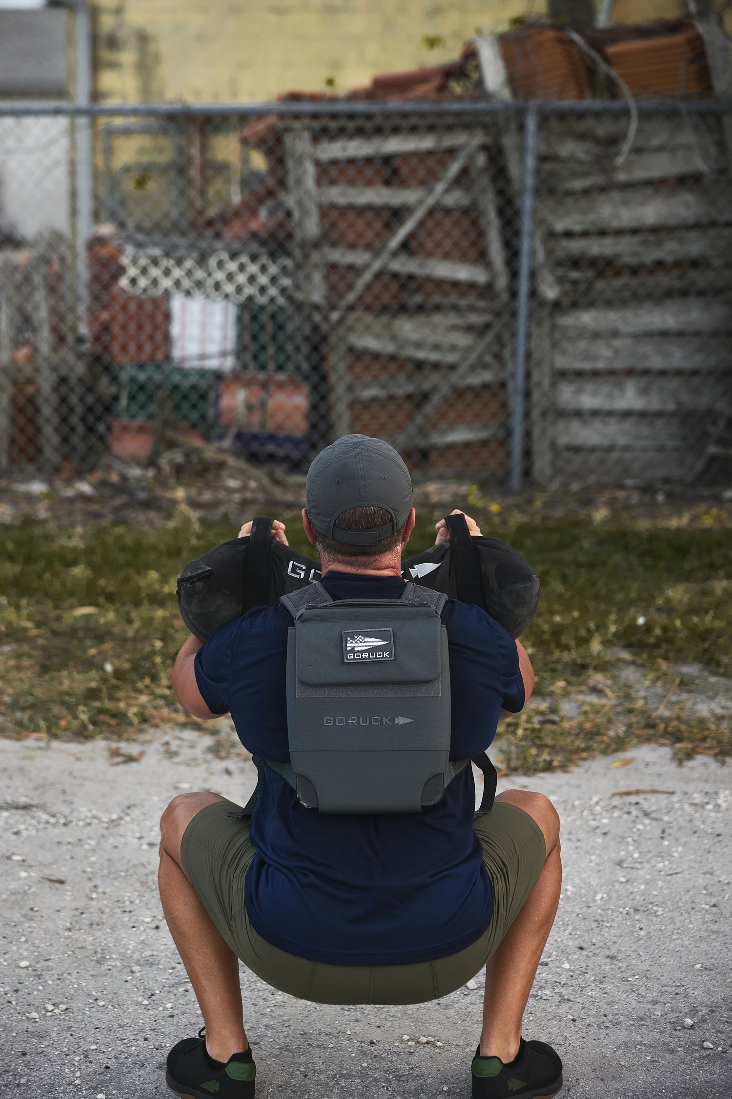 A man squats outdoors in front of a fenced area with stacked crates, wearing the RPC 3.0 and holding a 10lb Ruck Sandplate.