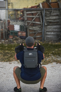 A man squats outdoors in front of a fenced area with stacked crates, wearing the RPC 3.0 and holding a 10lb Ruck Sandplate.