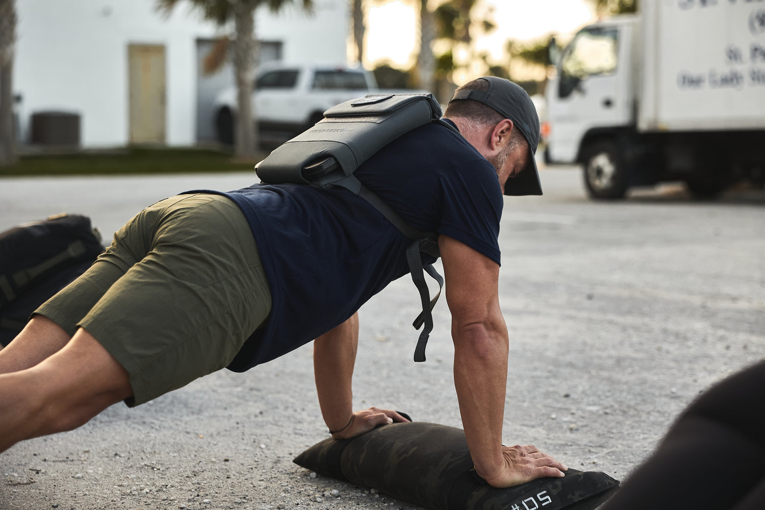 Man doing push-ups outdoors on a sandbag, wearing a cap, shorts, and the RPC 3.0 + 10lb Ruck Sandplate for added resistance.