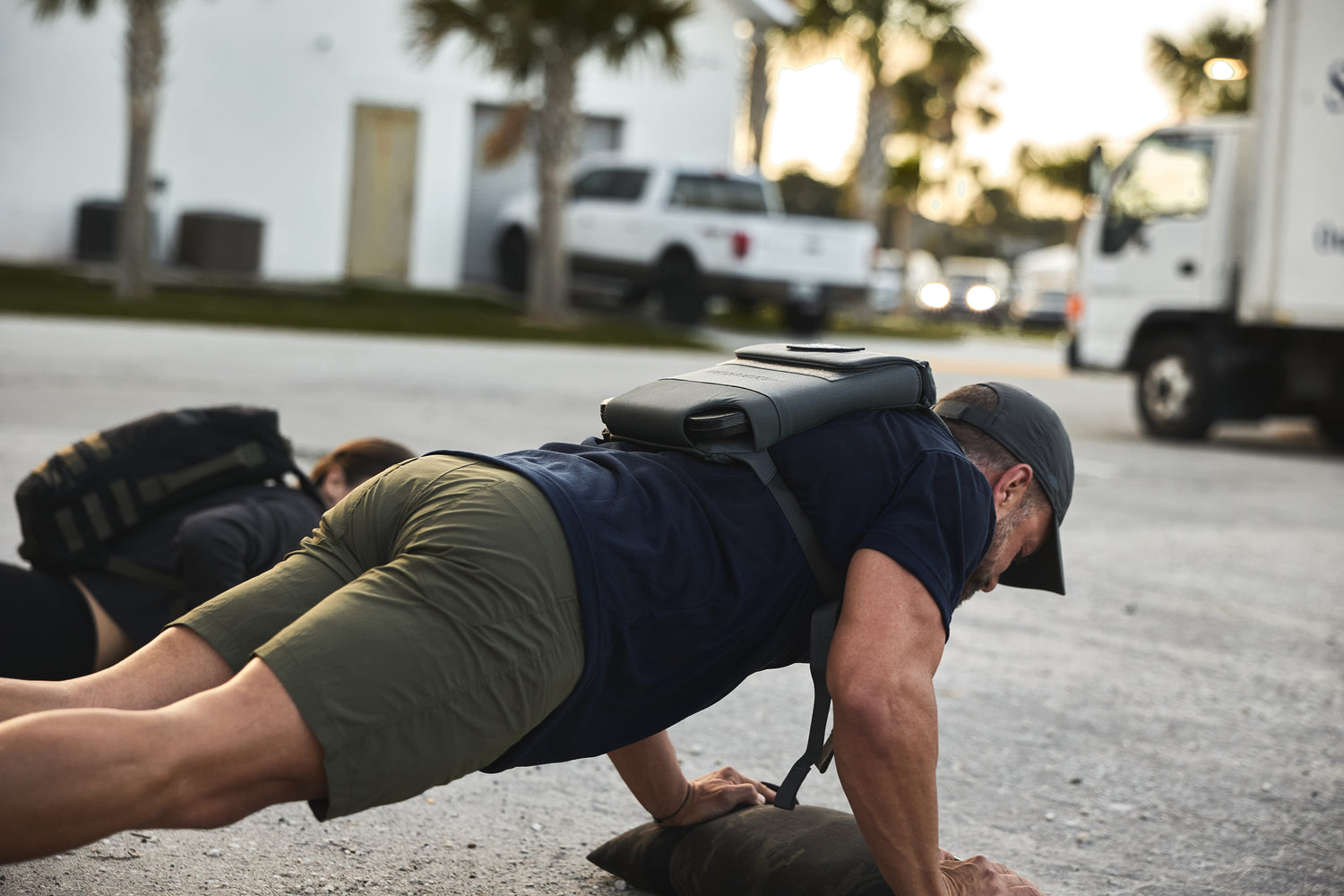 Man doing push-ups outdoors on gravel near a truck and palm trees, wearing the RPC 3.0 with a 10lb Ruck Sandplate.