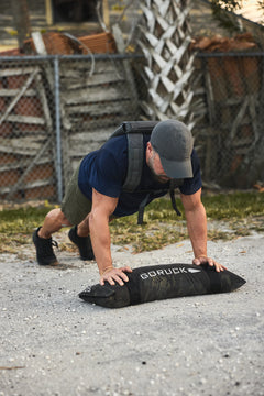 A man does push-ups outdoors on a sandbag, wearing a cap, shorts, T-shirt, and the RPC 3.0 + 10lb Ruck Sandplate for weighted training.