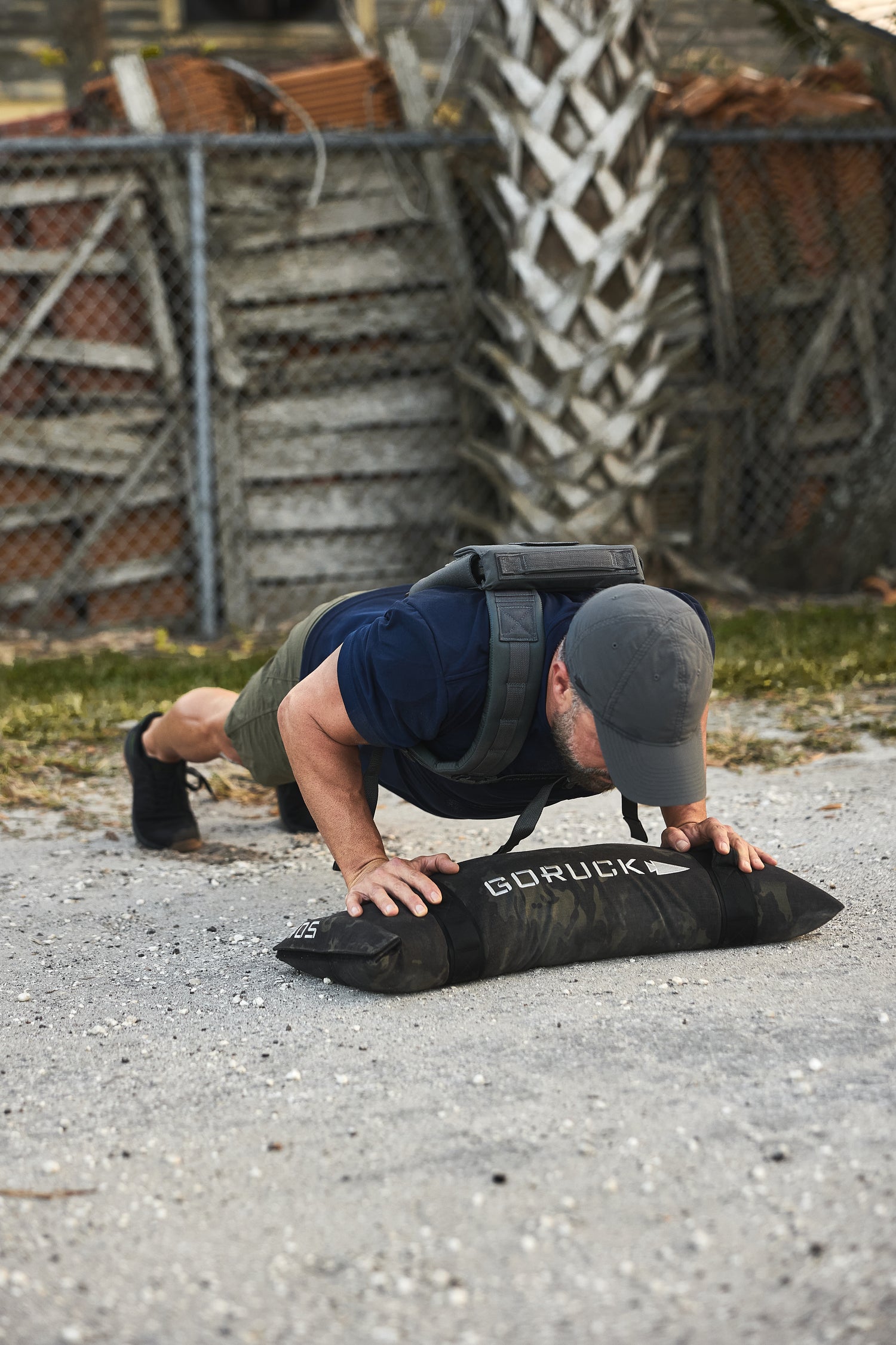Man performs push-ups outdoors on the RPC 3.0 + 10lb Ruck Sandplate, with a weighted vest, near a fence and palm tree.