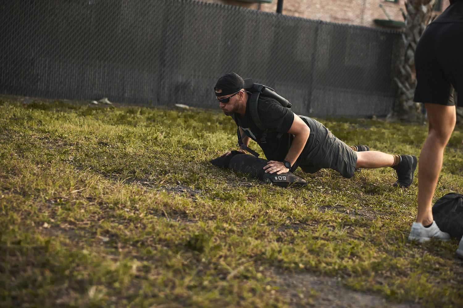 A man in black workout gear does push-ups on grass, wearing the RPC 3.0 + 10lb Ruck Sandplate for intense weighted training, with another person nearby.