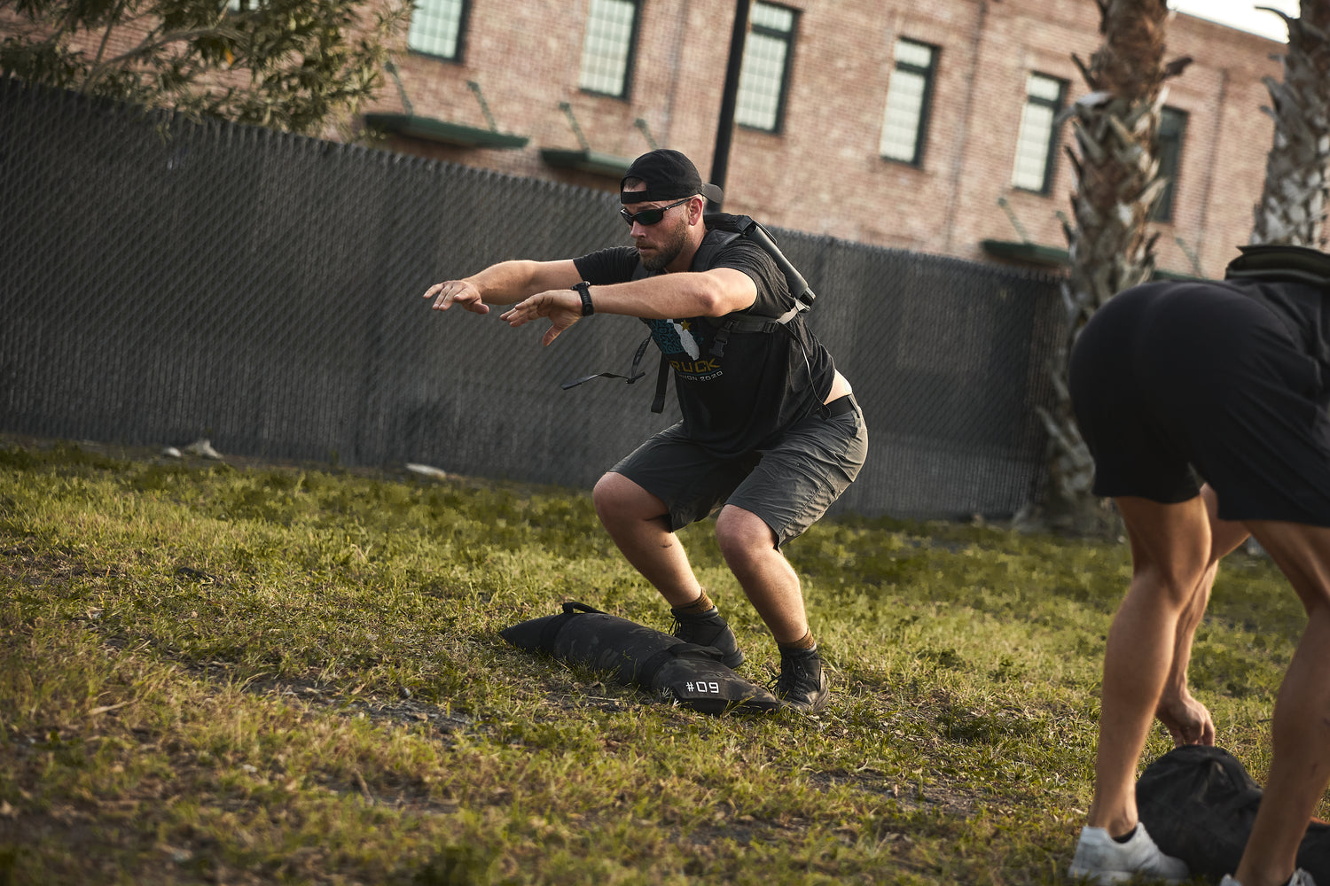 Man wearing the RPC 3.0 and sunglasses does squats outdoors, with a 10lb Ruck Sandplate on the ground in front of him and a cap on his head.