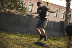 A man in athletic gear and cap jumps outdoors by a sandbag, wearing the RPC 3.0 + 10lb Ruck Sandplate vest, with a brick building in the background.