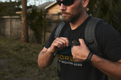 Wearing sunglasses and a dark GORUCK tee, a man fastens the chest strap on his backpack loaded with the RPC 3.0 + 10lb Ruck Sandplate, ready for an outdoor adventure.