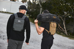 Two men walk outdoors doing weighted training—one with the RPC 3.0 + 10lb Ruck Sandplate and the other carrying a sandbag on his shoulder.