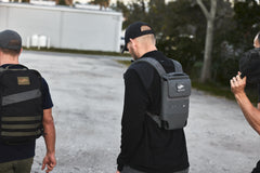 Three people with backpacks, hats, and the RPC 3.0 + 10lb Ruck Sandplate walk outdoors on a gravel surface, facing away from the camera.