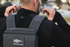 Man wearing the RPC 3.0 jacket and a backpack with a 10lb Ruck Sandplate, adjusting the straps outdoors.