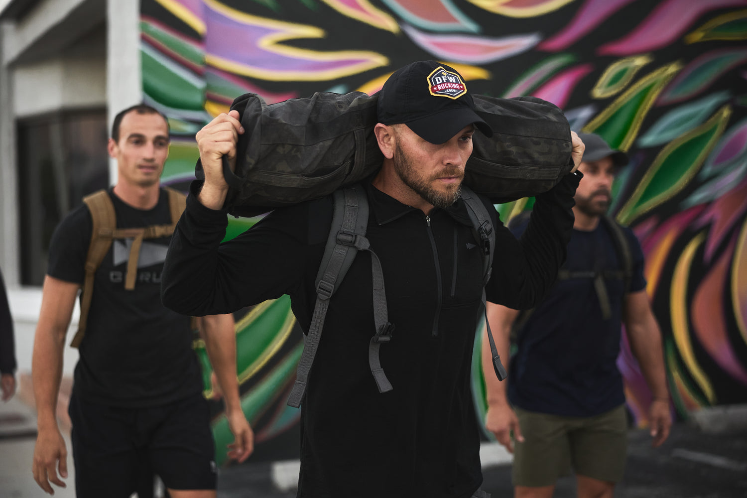 Three men exercise outdoors; one carries an RPC 3.0 + 10lb Ruck Sandplate on his shoulders in front of a colorful mural.