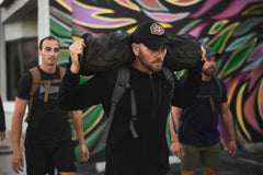 Three men exercise outdoors; one carries an RPC 3.0 + 10lb Ruck Sandplate on his shoulders in front of a colorful mural.