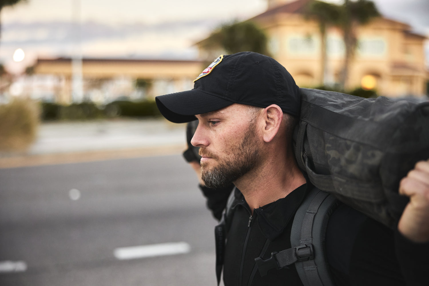 A man in a black cap walks outdoors carrying the RPC 3.0 backpack on his shoulders, weighted with a 10lb Ruck Sandplate.
