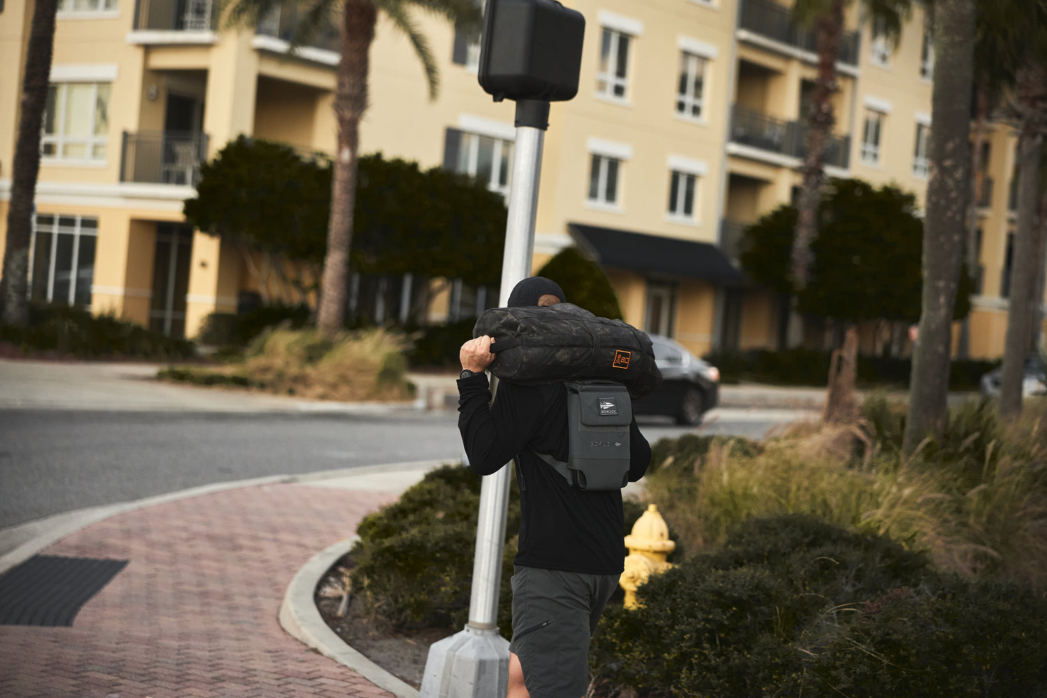 A person carries an RPC 3.0 + 10lb Ruck Sandplate and walks near an apartment building on a city street corner.