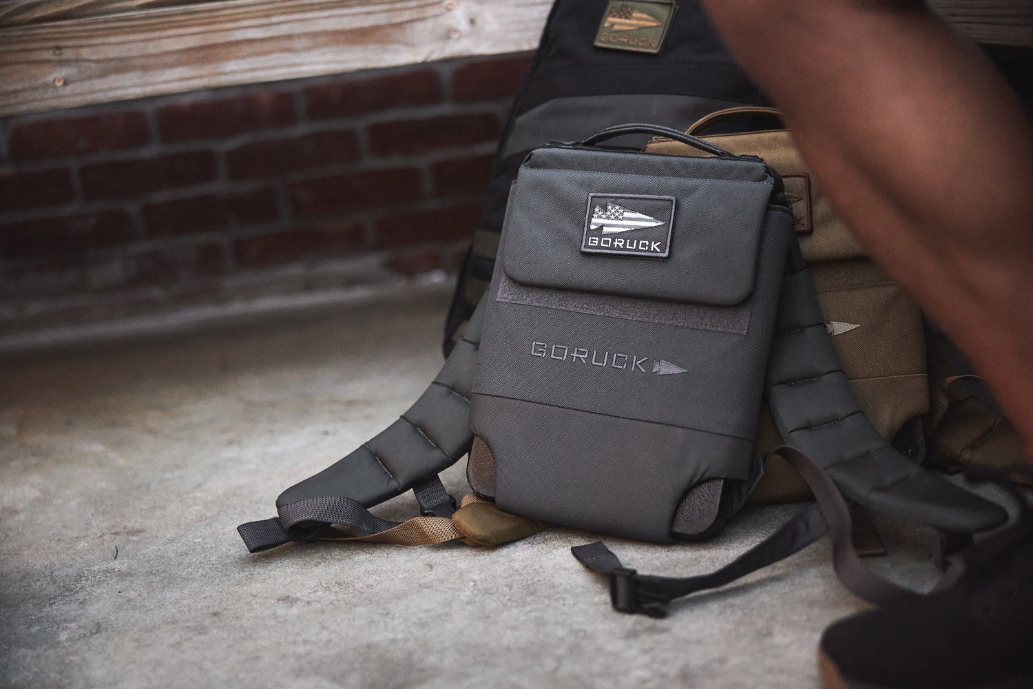 A gray GORUCK sling bag sits on the ground next to a tan backpack, someone’s leg, and the RPC 3.0 + 10lb Ruck Sandplate ready for action.