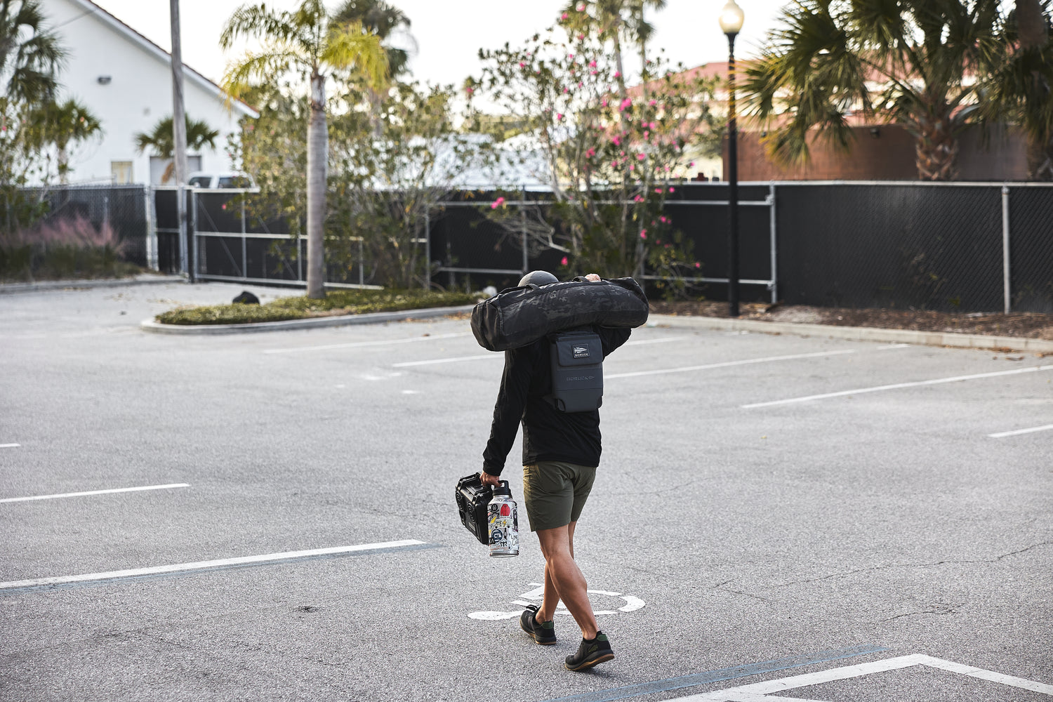 A person walks through a parking lot with palm trees, carrying a surfboard bag, helmet, and an RPC 3.0 + 10lb Ruck Sandplate.