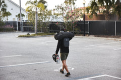 A person walks through a parking lot with palm trees, carrying a surfboard bag, helmet, and an RPC 3.0 + 10lb Ruck Sandplate.