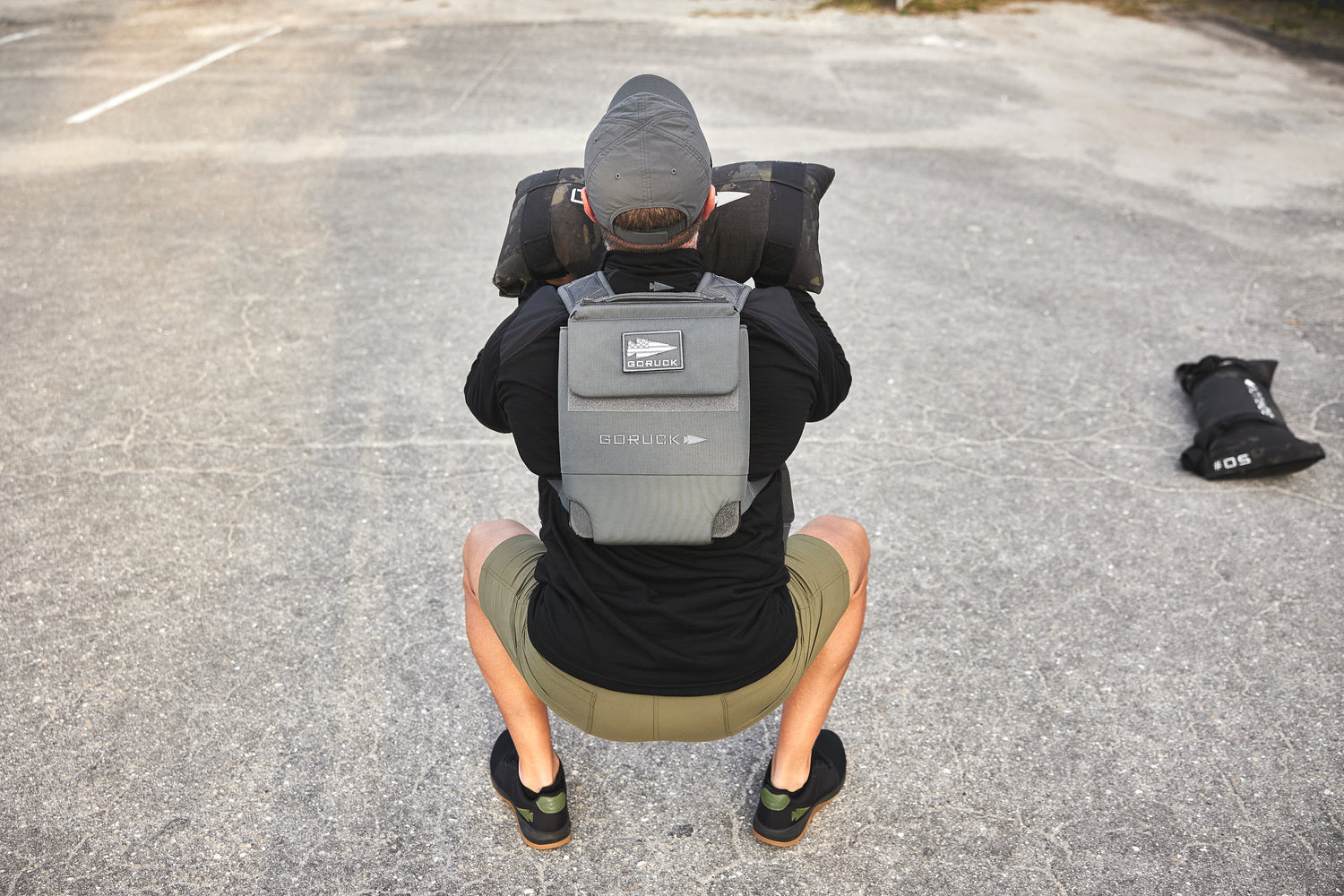 A person wearing the RPC 3.0 and lifting a 10lb Ruck Sandplate while squatting outdoors, with another sandplate on the ground nearby.