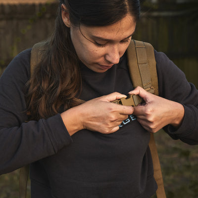 A woman fastens the chest strap of a Ruck Plate Carrier 3.0, which features padded shoulder straps, while standing outdoors.