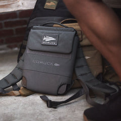 The Ruck Plate Carrier 3.0, a gray ballistic nylon backpack from GORUCK, sits on a concrete floor next to someone’s leg, resting against larger packs.