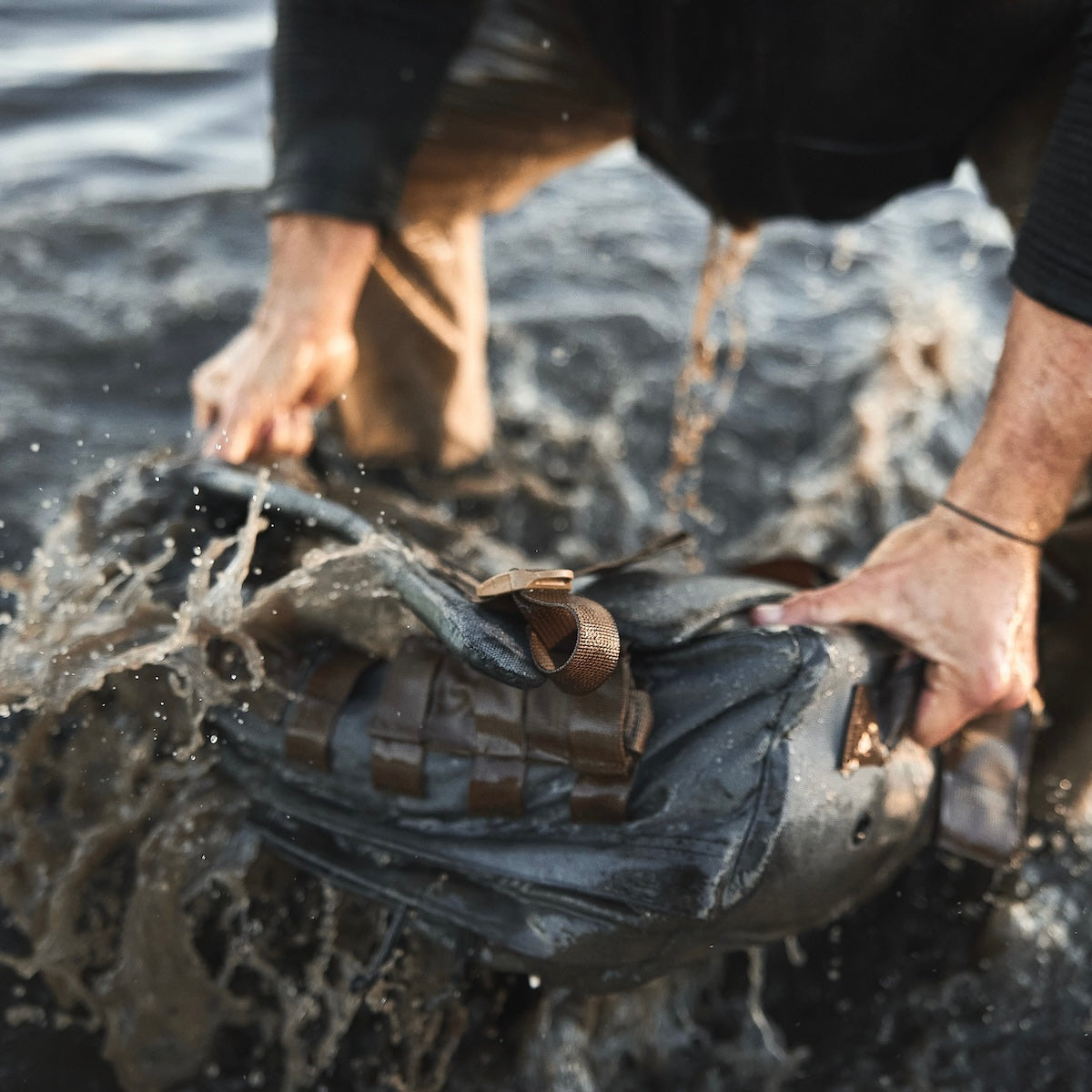 Person washing or dunking a rugged backpack in water, with splashes surrounding their hands and the bag.