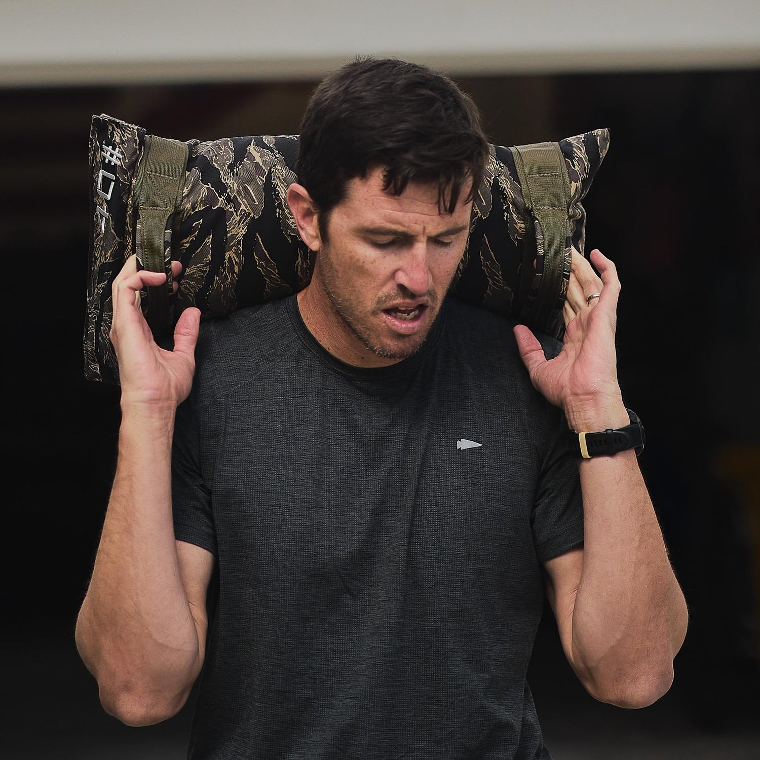 A man in a dark shirt lifts Simple Training Sandbags with a camo pattern onto his shoulders during an intense home workout.