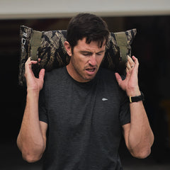 A man in a dark shirt lifts Simple Training Sandbags with a camo pattern onto his shoulders during an intense home workout.