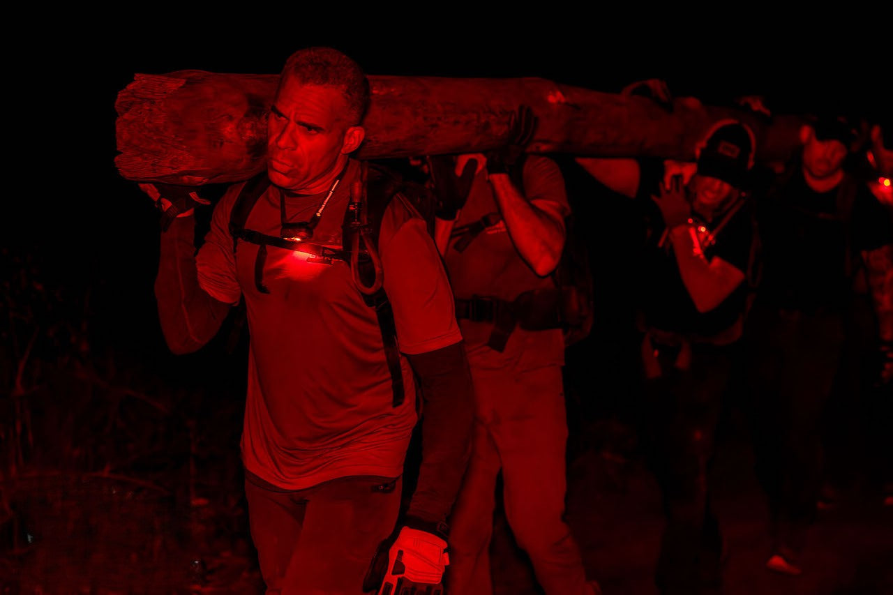 A group carrying a heavy log at night, illuminated by red headlamps and flashlights—evoking scenes from BRAGG 2027 training.