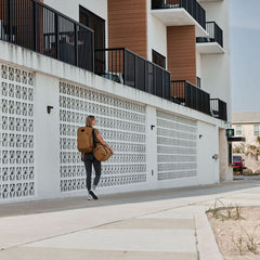 A person walks along a modern building's sidewalk, carrying a Kit Bag - X-PAC and a large brown X-PAC VX21 backpack reminiscent of Special Forces gear.