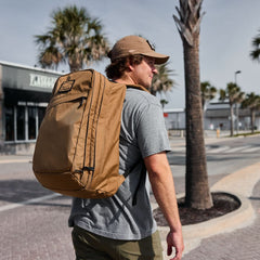 Man walking outdoors with tan GORUCK backpack and cap in sunny urban setting with palm trees