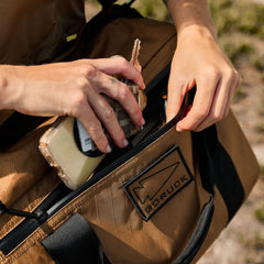 Close-up of hands placing a phone and keys into the brown Kit Bag - X-PAC, featuring a "GORUCK" patch and crafted from durable X-PAC VX21 material inspired by Special Forces gear.