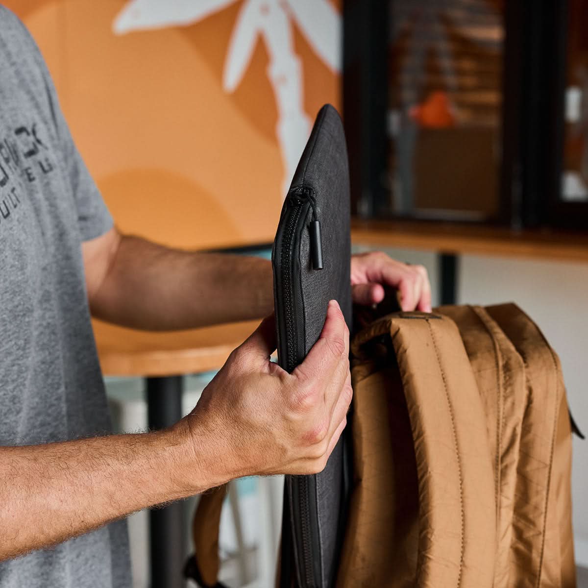 A person puts a lightweight black laptop sleeve into a tan GR2 X-PAC backpack indoors.