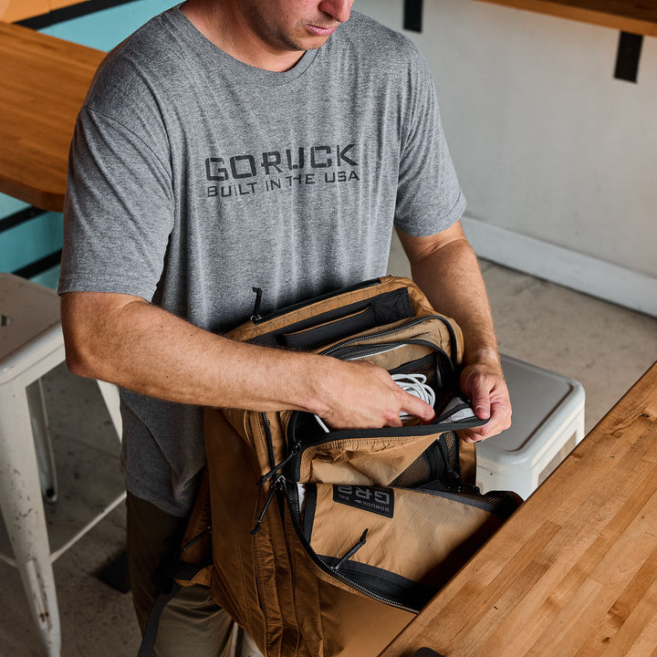 Person in a gray "GORUCK" shirt organizes cables inside a tan GR2 X-PAC travel backpack made of durable material at a wooden table.