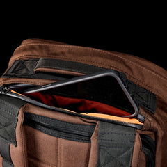 A tablet partially sticks out of the front zippered pocket of a GR1, a brown and black American-made backpack.