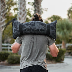 Outdoors, a person carries a Simple Training Sandbag in black camo on their shoulders, highlighting the versatility and essential role of training sandbags in any home gym.