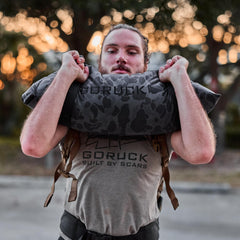 A person lifts a Simple Training Sandbag outdoors at sunset while wearing a GORUCK "Built by Scars" shirt, highlighting the versatility of sandbag workouts.