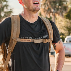 Man wearing a GORUCK shirt and backpack with MOLLE webbing, outfitted with the Training Sternum Strap, stands outdoors appearing tired and pauses to catch his breath.