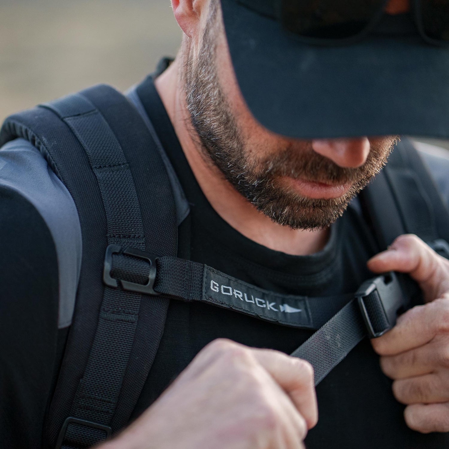 A man wearing a black cap adjusts the chest strap on a black Ruck Plate Carrier 3.0 with ergonomic lumbar support, outdoors.