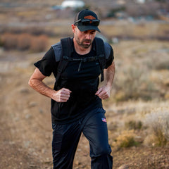 A man wearing Men’s Training Performance Joggers and a cap hikes on a dirt trail through a dry, open landscape.