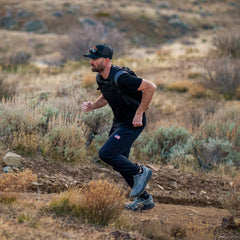 A man wearing Men’s Training Performance Joggers and a cap hikes uphill on a rocky, grassy trail in a dry, open landscape.