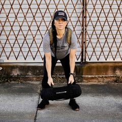 A person wearing a cap and gray t-shirt is training outdoors, holding a black Sandbags 2.0 from GORUCK, made with durable 1000D CORDURA. They stand confidently on a concrete surface in front of a metal gate, assured by the Scars Lifetime Guarantee.
