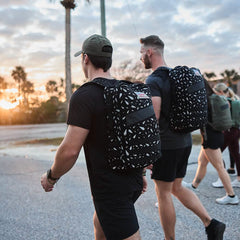 Two men with Rucker 4.0 - Reflective Spearhead rucksacks walk outdoors at sunset, joined by a group of people.